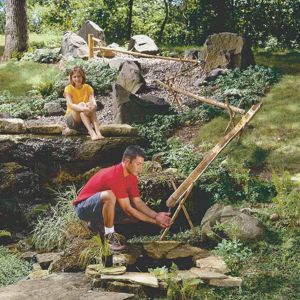 A man crouches by a small stream, adjusting a bamboo water feature, while a woman sits on stones nearby, surrounded by greenery and rocks.