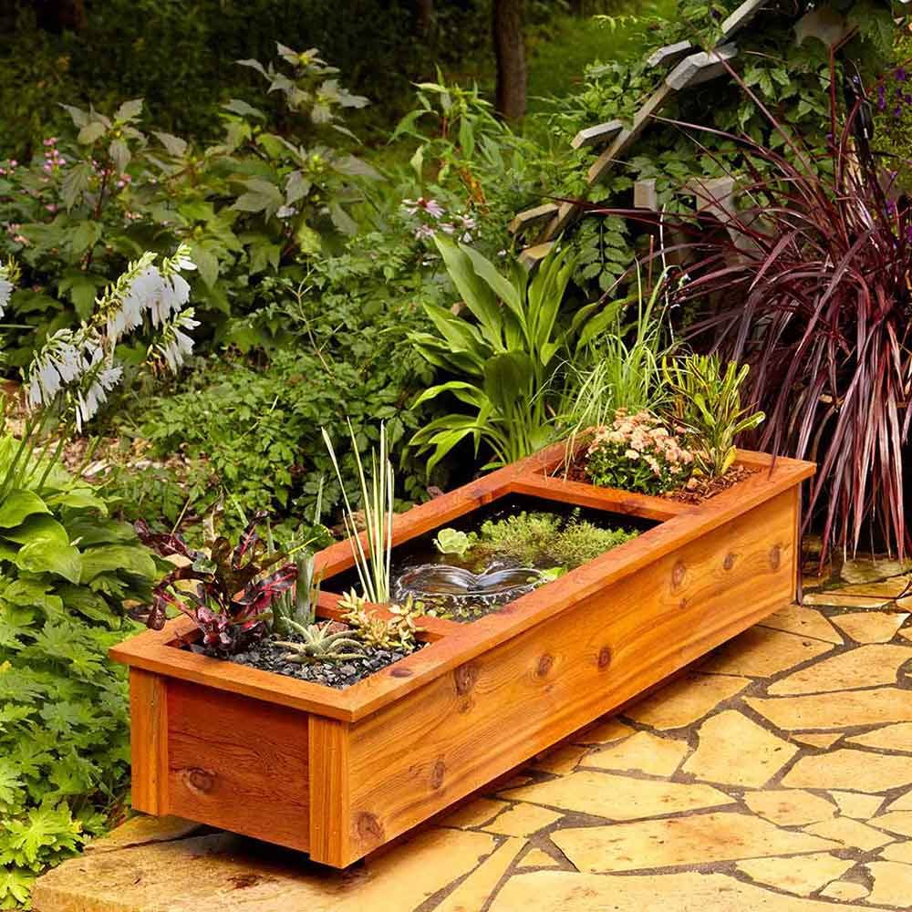 A wooden planter contains various plants and a small water feature, situated on stone paving amidst lush greenery and vibrant foliage.
