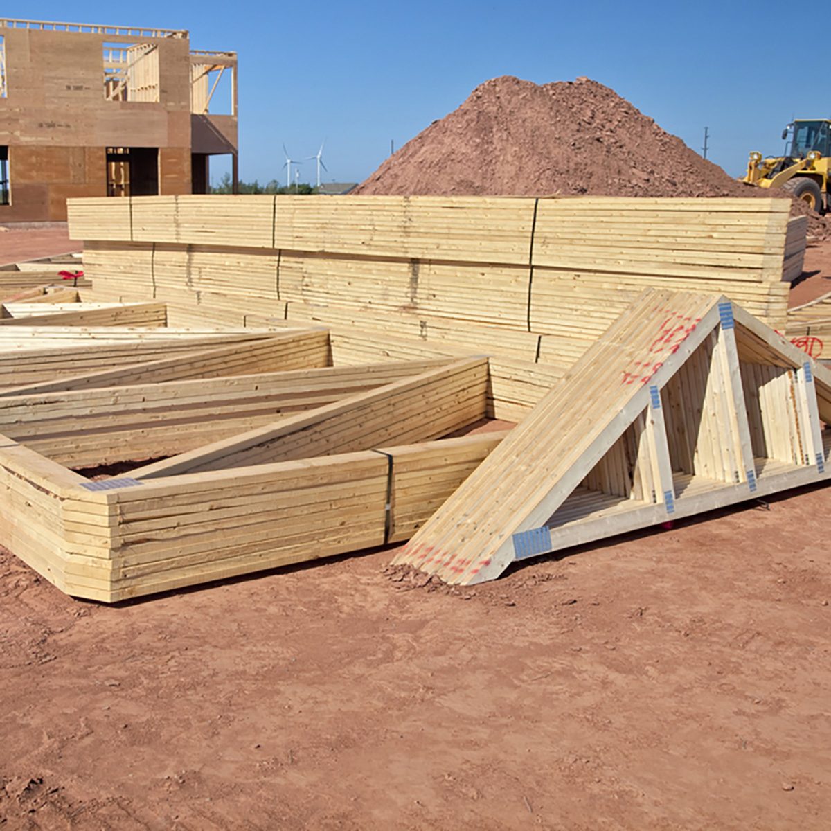 Stacked wooden beams and trusses are arranged on a construction site with a backdrop of clear blue skies and a dirt mound in the distance.