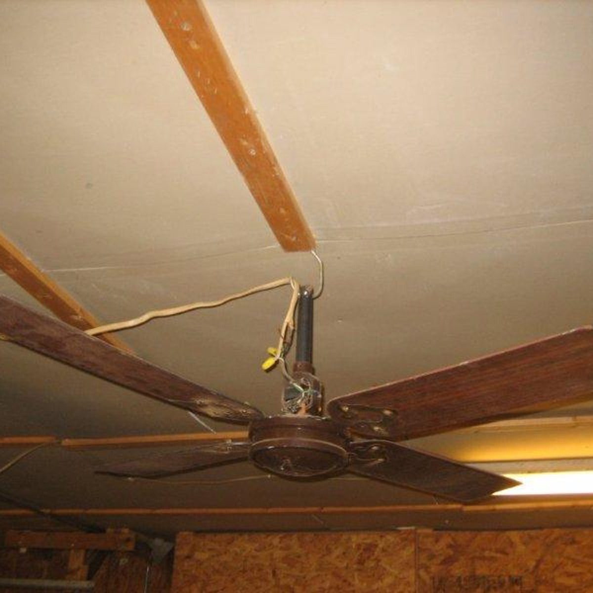 A ceiling fan hangs from a wooden beam, with exposed wiring and a yellow clip, inside a dimly lit room with bare walls.