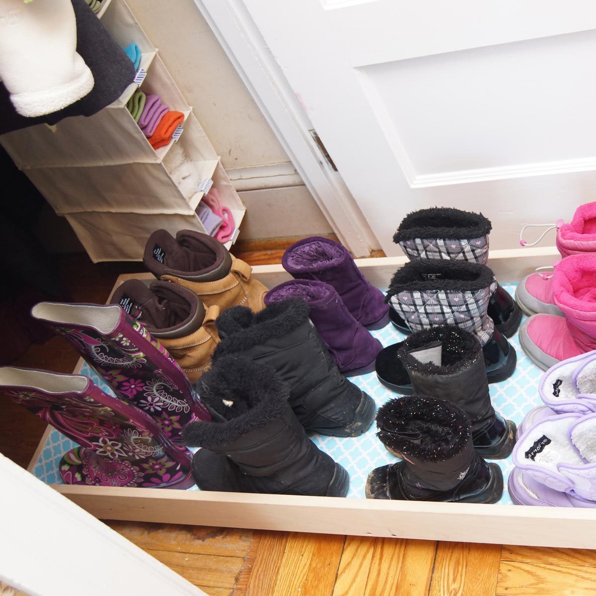 A variety of winter boots are neatly arranged on a storage tray by a door, with colorful socks visible in the background.