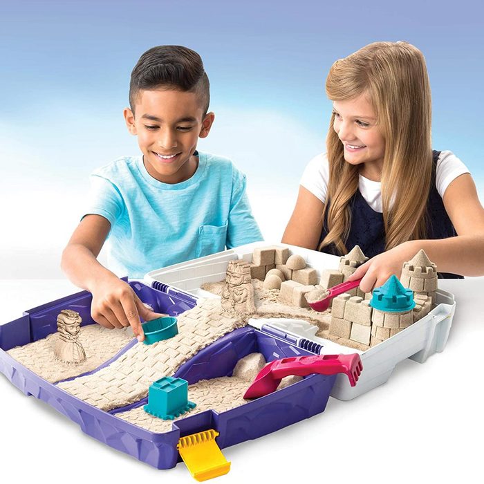 Two children are playing with kinetic sand, creating structures and shapes in a divided tray, smiling as they interact in a bright, playful setting.