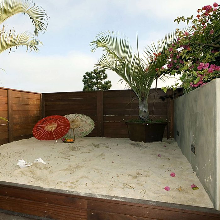 A sandbox features two decorative umbrellas, a potted palm, and scattered flowers, enclosed by wooden fencing and a concrete wall, under a cloudy sky.