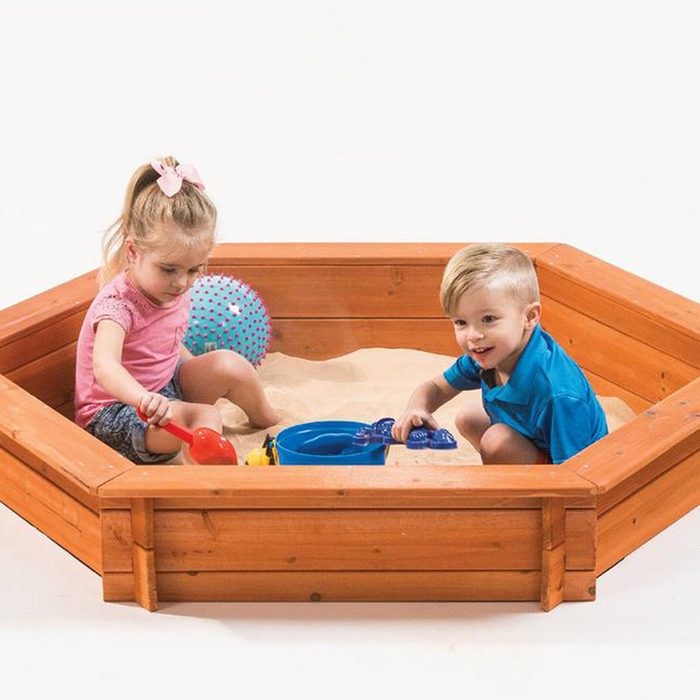 Two children play in a wooden sandbox, one using a red shovel and the other holding a blue toy, surrounded by sand and a beach ball.
