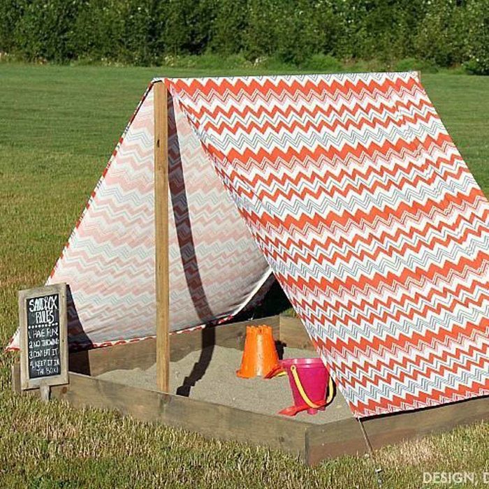 A patterned tent structure provides shade over a small sand area, with colorful buckets resting inside. It sits on a grassy field.