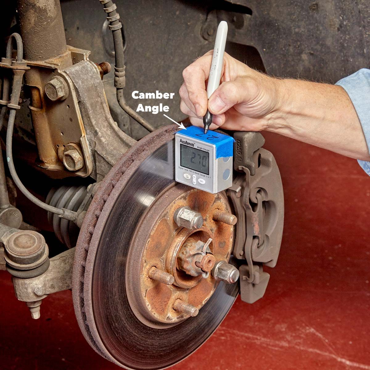 A person measures the camber angle of a vehicle's brake rotor using a digital gauge on a red workshop floor.
