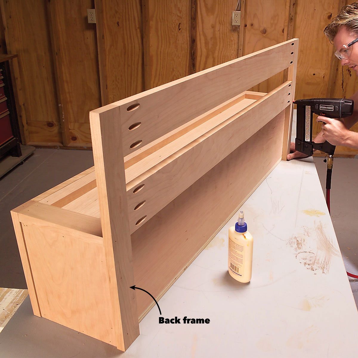 A wooden frame is being assembled with nails, while glue sits nearby on a workbench in a woodshop surrounded by wooden walls.