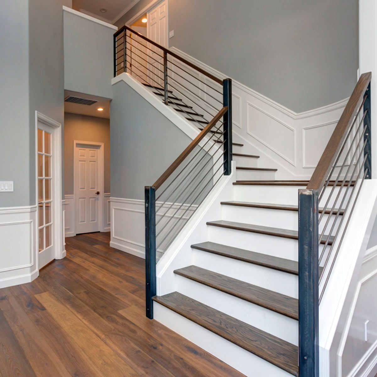 A wooden staircase curves upward, bordered by sleek railings, in a spacious hallway with gray walls and polished wooden flooring leading to a closed door.
