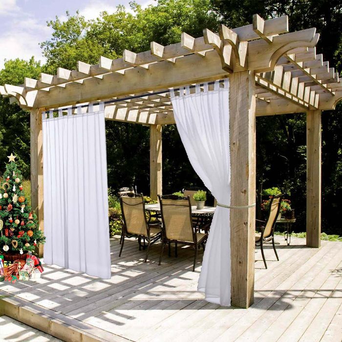 A wooden pavilion features white curtains, surrounding a table with chairs. A small Christmas tree stands nearby, amidst greenery and sunlight.