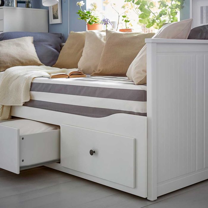 A white daybed with striped mattress rests against a wall, accompanied by pillows and a blanket. A book lies open on the bed.