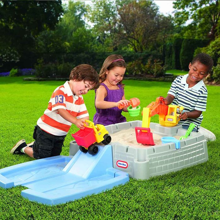 Three children play with toy trucks and shovels in a sandpit on a grassy lawn, enjoying a sunny day outdoors.