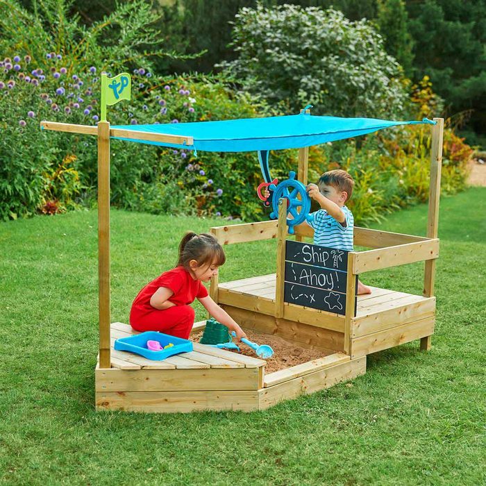 Children play in a wooden sandbox designed like a ship, using toys while a blue canopy provides shade in a garden setting.