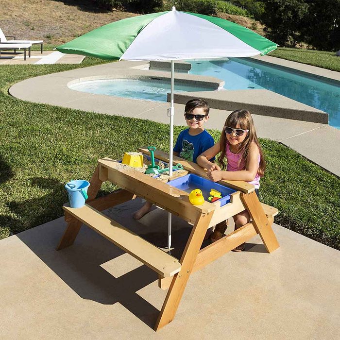 A wooden sand and water table under a green and white umbrella; two children are playing with toys beside a swimming pool on a sunny day.