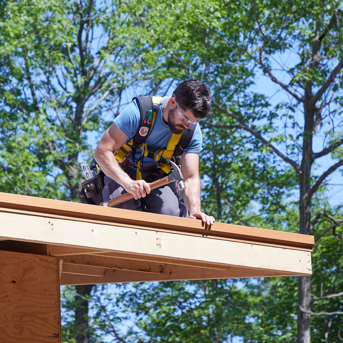 A man in safety gear hammers a piece of wood while standing on a roof, surrounded by green trees under a clear blue sky.