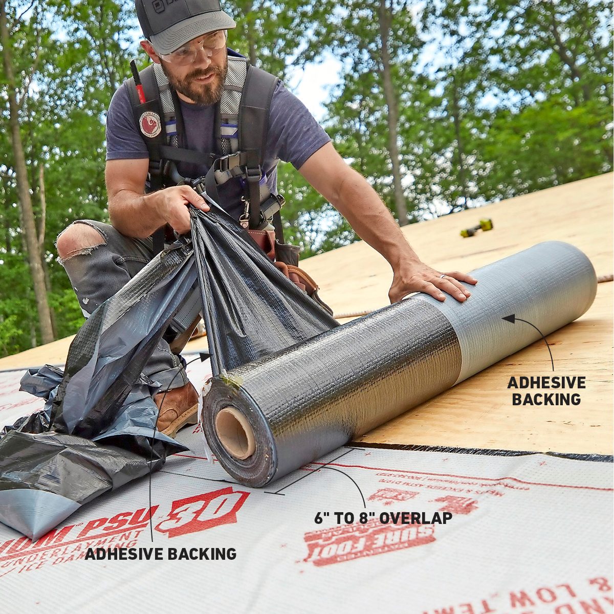 A man unrolls a black material with adhesive backing on a wooden roof, preparing for installation amidst a green, wooded environment.