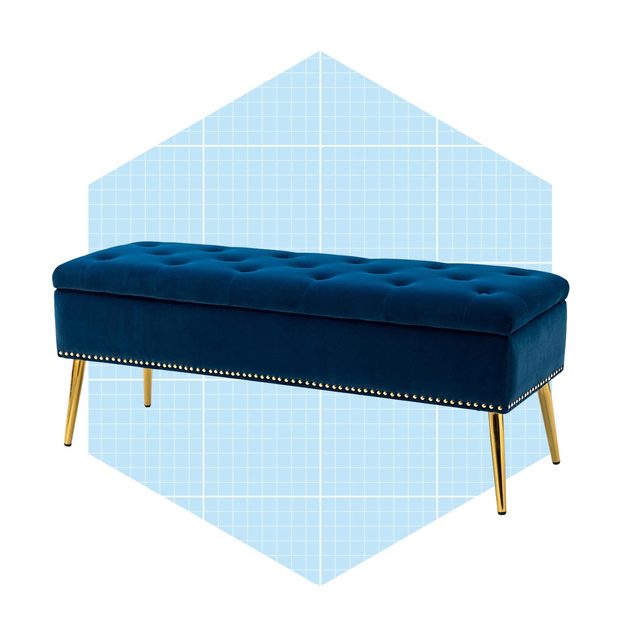 A blue velvet bench with golden legs rests against a light blue geometric background, featuring tufted upholstery and decorative nailhead trim.
