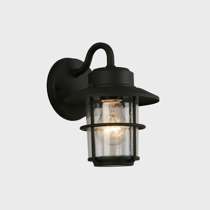 A wall-mounted lantern emits warm light, featuring a glass enclosure and a black metal framework, set against a neutral background.