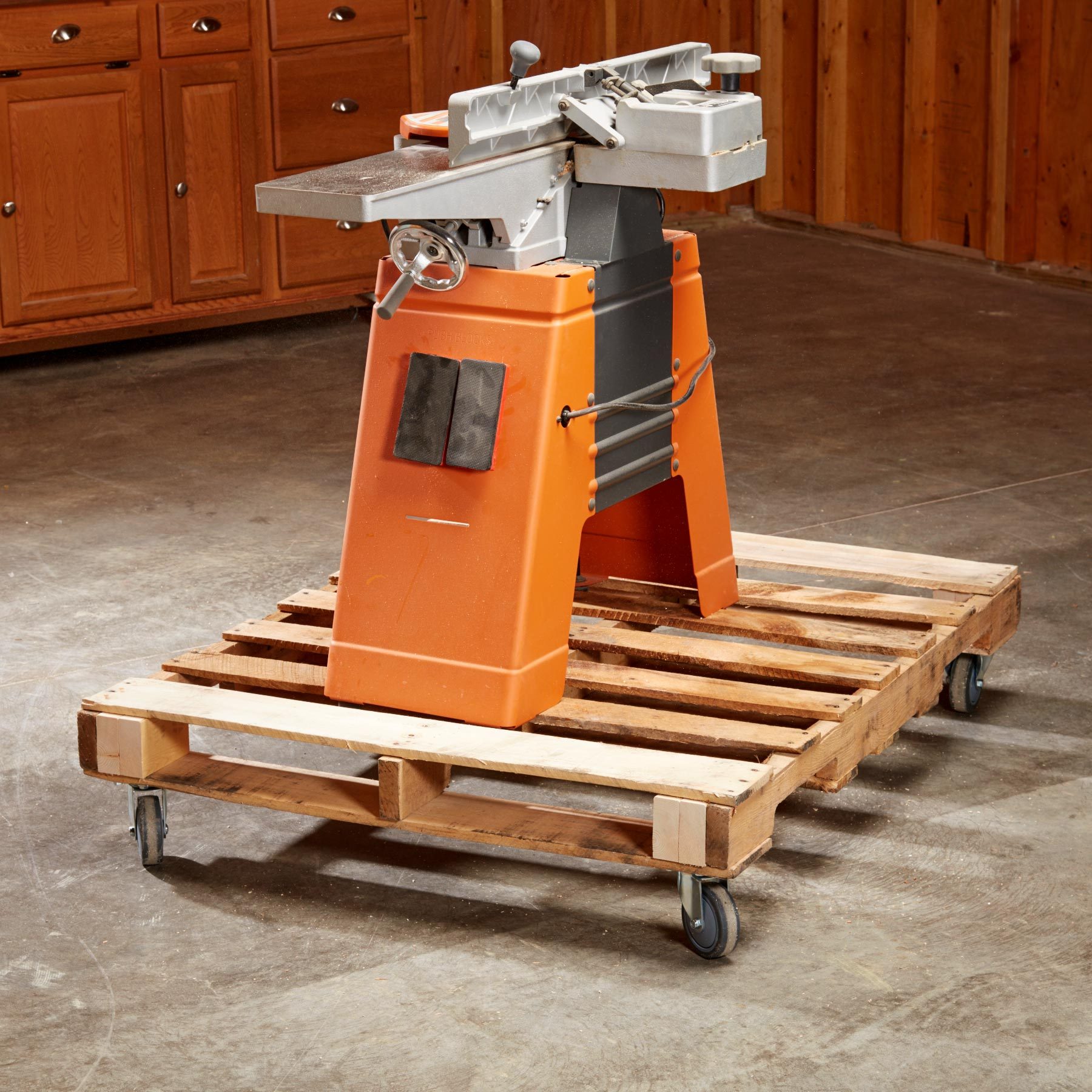 An orange woodworking machine sits on a wooden pallet with wheels, positioned in a room with wooden cabinetry and a concrete floor.