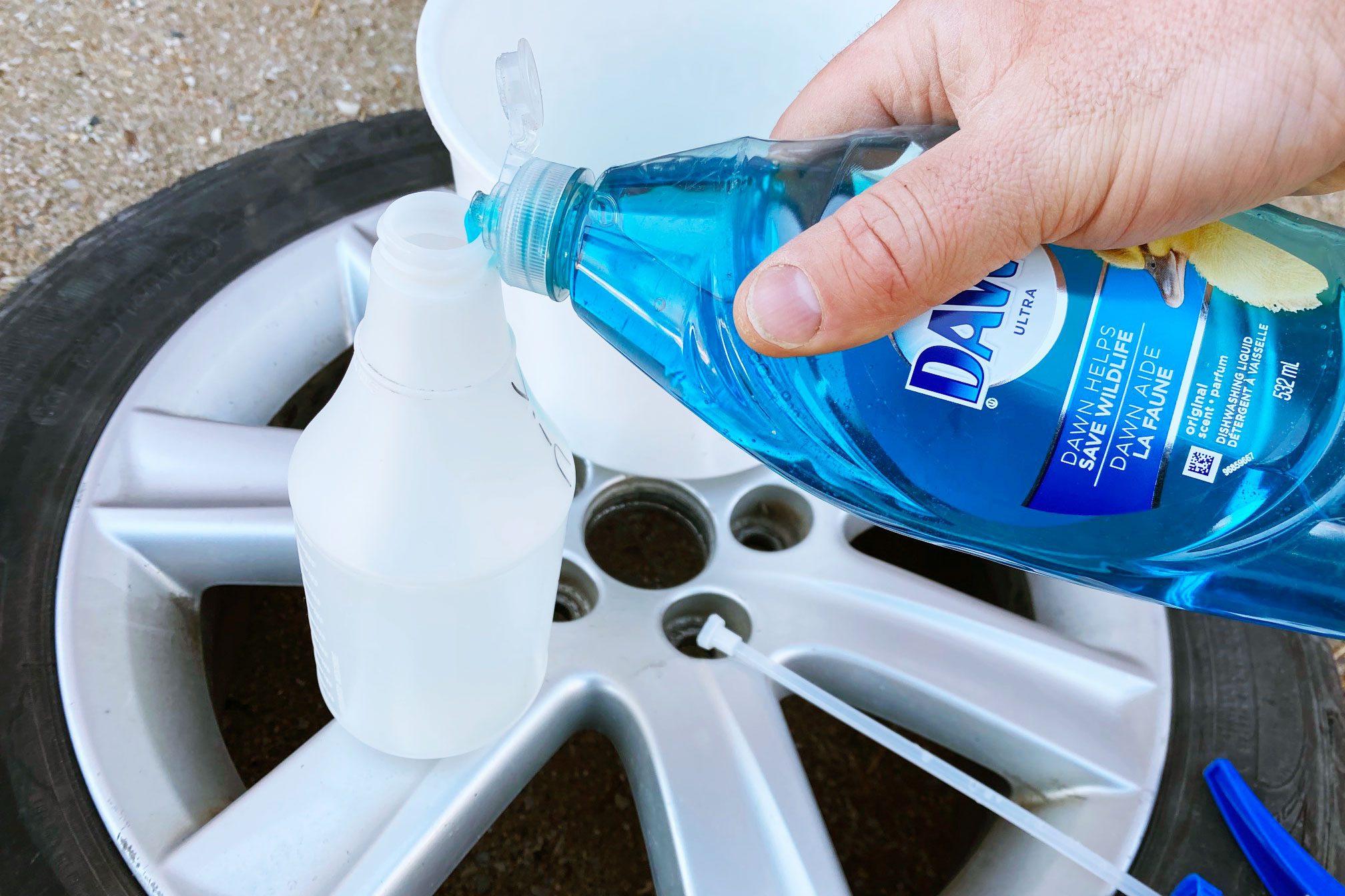 A hand pours blue dish soap into a clear bottle beside a wheel and tire on a gravel surface. A white bucket is in the background.