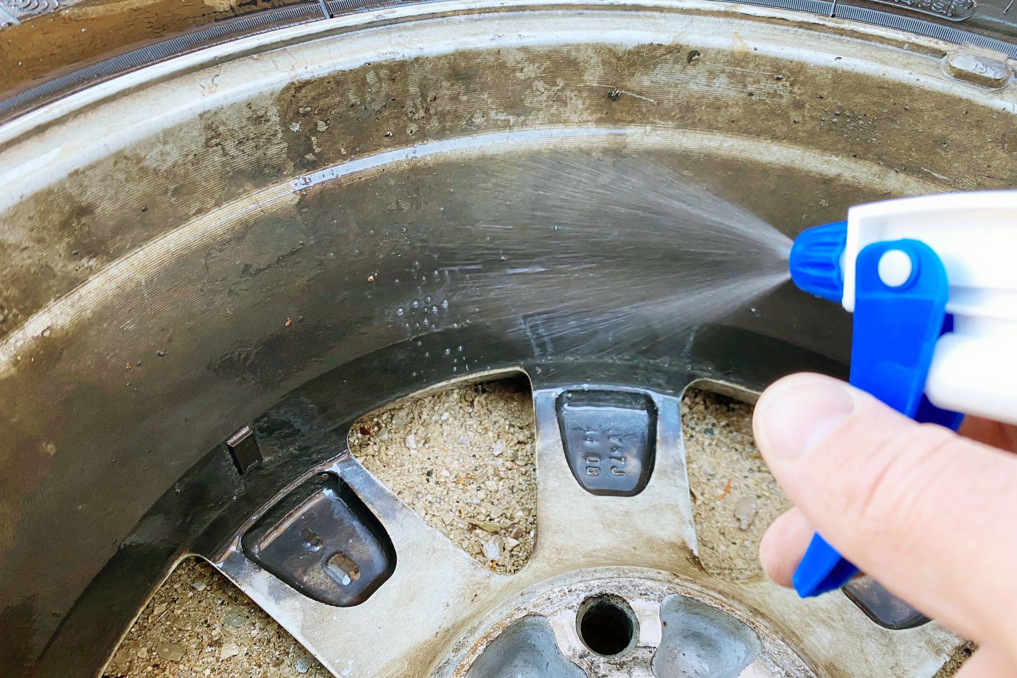 A hand sprays cleaner on a dirty car rim, with sandy ground visible in the background, targeting areas needing maintenance and cleaning.
