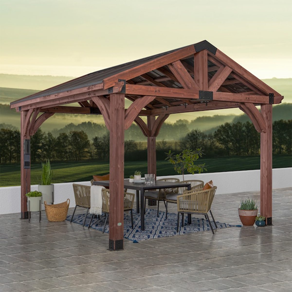 A wooden gazebo shelters a dining table and chairs, surrounded by potted plants, on a tiled patio with a scenic, misty landscape in the background.