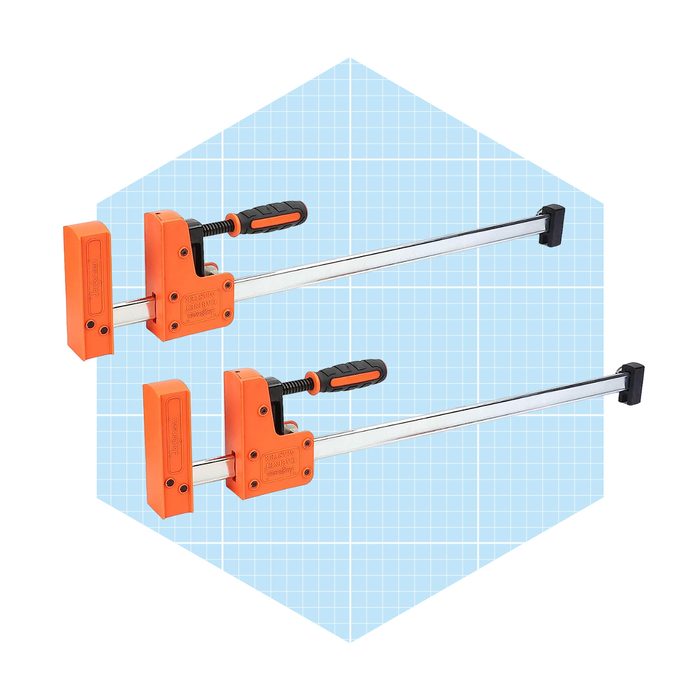 Two orange clamps with silver metal bars are positioned side by side, featuring black rubber grips, set against a light blue grid background.